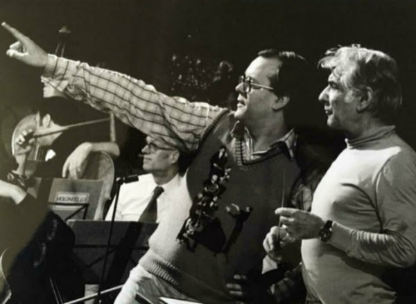 Black-and-white photo of two men standing side by side on a stage rehearsal space; the man at center (Humphrey Burton), wearing glasses, a vest, and a checkered shirt, points off to the left, while the man beside him (Leonard Bernstein), in a light-colored sweater, looks in the same direction holding a baton.