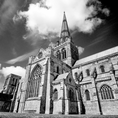 The image showcases a black and white photograph of Chichester Cathedral, a historic Anglican cathedral located in Chichester, England. The cathedral exhibits classic Gothic architectural features, including pointed arches, intricate tracery, and towering spires. The central spire, a striking feature of the structure, rises into a partly cloudy sky, creating a dramatic and awe-inspiring backdrop. The photograph is taken from a low angle, emphasizing the cathedral's grandeur and height. 