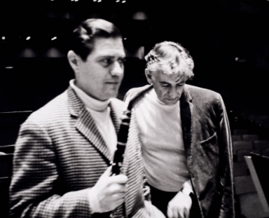 Man with Clarinet stands next to conductor pointing at score on a music stand.