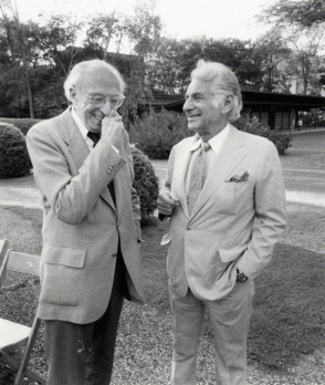 A black and white image of Aaron Copland (left) and Leonard Bernstein right standing outdoors on the grounds of Tanglewood, both in suits. Copland is laughing with his hand partially covering his mouth, and Bernstein is smiling. 