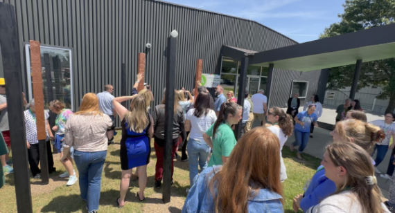 Students and teachers walk around outdoor art instalation, touching the raised relief designs on copper-covered poles.
