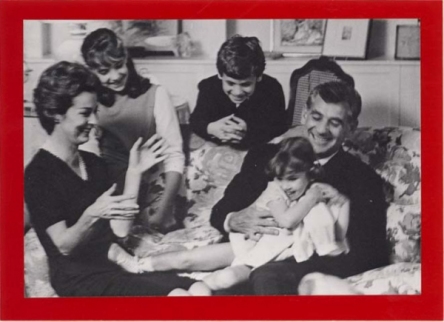 Family consisting of wife, husband, two daughters, and one son laughing on a couch.